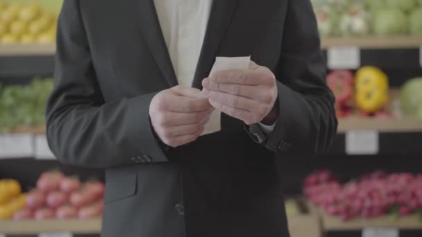 Vista frontal de un hombre caucásico irreconocible sosteniendo la factura en las manos y comprobando el precio de los alimentos comprados en la tienda de comestibles. Un tipo serio examinando el costo de las compras en el supermercado. Estilo de vida, finanzas. S-log 2 . — Vídeos de Stock