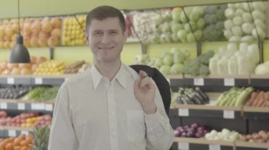 Kahverengi gözlü, markette poz veren neşeli, beyaz, esmer bir adamın portresi. Süpermarkette kameraya gülümseyen beyaz tişörtlü mutlu yetişkin adam. Neşe, yaşam tarzı, tüketim konsepti. S-log 2.