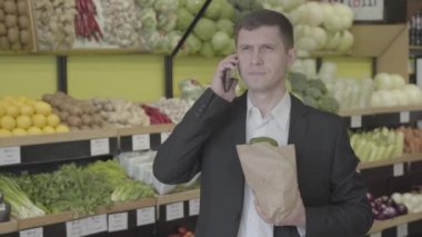 Markette telefonla konuşan esmer, kahverengi gözlü, beyaz bir adamın portresi. Yetişkin, kendine güveni tam bir adam elinde torbayla akıllı telefondan sohbet ediyor. Alışveriş, yaşam tarzı, sağlıklı beslenme. S-log 2.