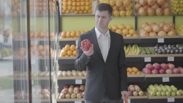 Porträtt av självsäker vit man undersöker röd paprika, sträcker den till kameran och ler. Glada kunder väljer grönsaker i mataffären. Livsstil, shopping, hälsosam mat. S-log 2 — Stockvideo
