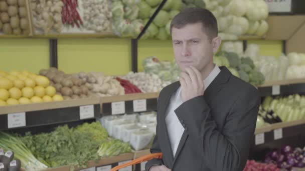 Retrato de morena caucasiano homem com olhos castanhos em pé no supermercado e pensamento. Cara séria em terno elegante decidir sobre variedade para comprar. Estilo de vida, consumismo, compras, varejo. S-log 2 . — Vídeo de Stock