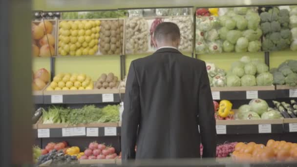 Bakåt syn på unga kaukasiska mannen i kostym tittar igenom hyllor med frukt och grönsaker i livsmedel och tänkande. Vuxen kille som väljer varor i butik. Konsumtion, livsstil. S-log 2. — Stockvideo