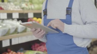 Raflarda yürüyüp tablet kullanan üniformalı tanınmamış bir erkek bakkal. Perakende dükkanındaki sebze ve meyve listesini kontrol eden yetişkin bir adam. Ticaret, tüketim, yaşam tarzı. S-log 2.