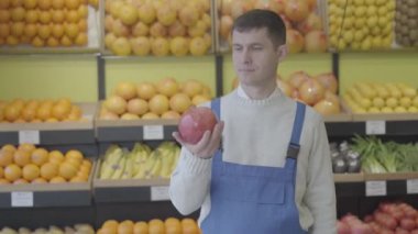 Elinde nar tutan ve kameraya baş parmağını gösteren beyaz üniformalı esmer adamın portresi. Bakkalda durup gülümseyen kendine güvenen bir işçi. Ticaret, sağlıklı beslenme. S-log 2.