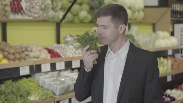 Retrato del hombre caucásico sonriente oliendo a fresco manojo de vegetación en la tienda de comestibles. Un joven con traje seleccionando eneldo en el supermercado. Hierbas, especias, cocina, estilo de vida. S-log 2 . — Vídeos de Stock