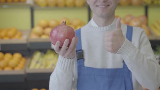 Alegre hombre caucásico desconocido en bata azul sosteniendo granada en la mano y mostrando el pulgar hacia arriba. Trabajador feliz posando en el supermercado con jugosa fruta fresca. Alimentación saludable, dieta, comercio. S-log 2 . — Vídeo de stock