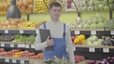 Bakkalda klasör ve patlıcanla poz veren genç, beyaz bir çalışan. Süpermarkette çalışan üniformalı gülümseyen adamın portresi. İş, ticaret, meslek, yaşam tarzı. S-log 2.