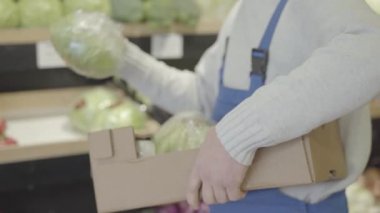 Tanınmayan bir erkek çalışanın marketteki raflara lahana koyup gitmesi. Üniformalı adam süpermarkette sebze çeşitleri ekliyor. Ticaret, perakende, iş, hizmet. S-log 2.