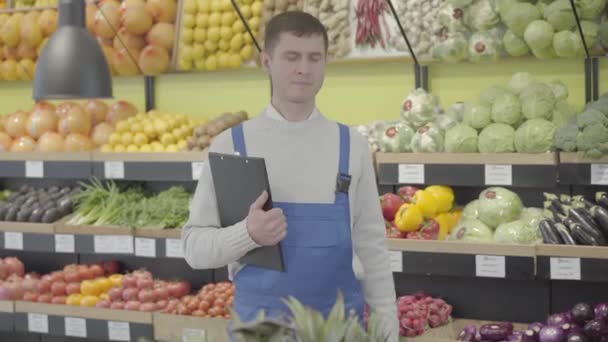 Ung positiv kaukasisk anställd poserar i matvaror med mapp och aubergine. Porträtt av en leende man i uniform som arbetar i snabbköpet. Affärer, handel, yrke, livsstil. S-log 2. — Stockvideo