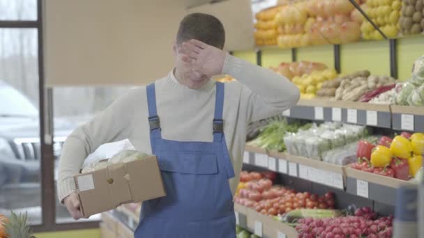 Cansado homem caucasiano limpando a testa com a mão, suspirando e saindo com caixa de couves. Retrato de empregado de mercearia exausto que trabalha no supermercado. Estilo de vida, retalho, comércio. S-log 2 . — Vídeo de Stock