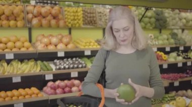Bakkaldan meyve alan mutlu, kendine güvenen beyaz kadın portresi. Yaşlı sarışın ev hanımı sepete pomelo koyup gülümsüyor. Zevk, yaşam tarzı, alışveriş, tüketim. S-log 2.
