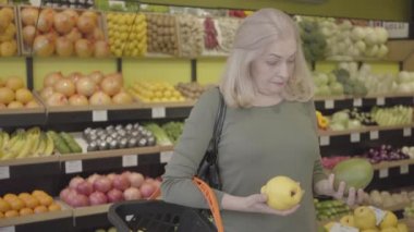 Yaşlı, kendine güvenen beyaz kadının portresi. Markette armut ve armut arasında seçim yapıyor ve her iki meyveyi de almaya karar veriyor. Ciddi sarışın kadın süpermarketten sağlıklı yiyecekler alıyor. Tüketim. S-log 2.