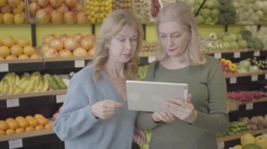 Markette tablet kullanan Kafkasyalı yaşlı kadınların portresi. İki ev hanımı mal ile konuşma arasında duruyor. Kablosuz teknoloji, yaşam tarzı, tüketim, alışveriş. S-log 2.