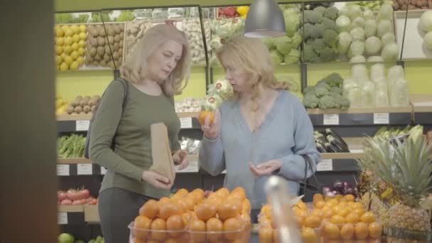 Zwei erwachsene kaukasische Hausfrauen pflücken Mandarinen im Supermarkt. Ernsthafte Seniorinnen legen Früchte in Papierschachteln und unterhalten sich. Lebensstil, Vitamindiät, Vegetarismus, Konsum. S-Log 2. — Stockvideo