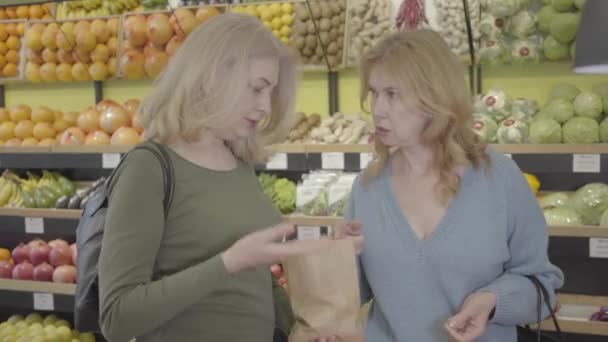 食料品店での購入について議論している2人の自信に満ちた白人主婦の肖像。小売店の商品の列と話の間に立っているシニアブロンドの女性.生活様式消費主義。S-log 2. — ストック動画