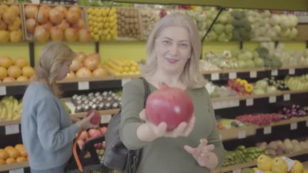 Fröhliche ältere kaukasische Frau streckt Granatapfel in die Kamera und redet, während eine andere Kundin im Hintergrund Früchte riecht. Porträt einer freudigen Kundin, die Lebensmittel im Lebensmittelgeschäft kauft. S-Log 2. — Stockvideo