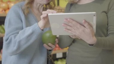 İki tanınmayan Kafkasyalı kadın, marketteki pomelo köklerini kontrol etmek için tablet kullanıyor. Ciddi bayanlar süpermarketten alışveriş yapıyor. Sağlıklı yiyecekler, yaşam tarzı, tüketim, diyet. S-log 2.