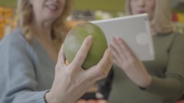 Arka planda tablet kullanan bulanık kadınlar gibi beyaz kadın ellerinin pomelo tutuş görüntüsü. Kendinden emin yaşlı hanımlar, modern teknolojilerin kullanıldığı meyvenin kökenini doğruluyor. S-log 2.