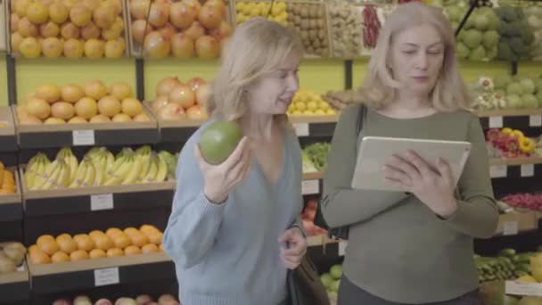 Впевнена в собі блондинка-кавказька жінка з табличкою, яка радить другові вибрати кращий фрукт у продуктовій промисловості. Позитивна зріла жінка-друзі купують соковиту органічну їжу в супермаркеті. S-log 2. — стокове відео