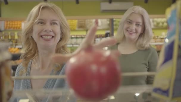 食料品店の棚に寝そべっている新鮮な有機ザクロのジューシーな新鮮な有機ザクロから、買い物かごに入れる2人のシニア白人女性の幸せな顔に焦点を当てます。生活様式消費主義。S-log 2. — ストック動画