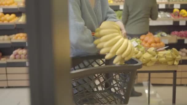 Mujer caucásica adulta irreconocible con cesta de la compra caminando por filas en la tienda de comestibles y tomando frutas. Cliente femenino comprando plátanos orgánicos y mandarinas. Estilo de vida, comida vegana. S-log 2 . — Vídeo de stock