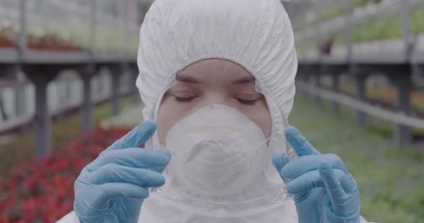 Close-up gezicht van vrouwelijke bioloog of wetenschapper die beschermende brillen opzet. Portret van een jonge blanke vrouw in een masker en een wit pak poserend in een kas. Tuinieren. Cinema 4k ProRes Hoofdkwartier. — Stockvideo