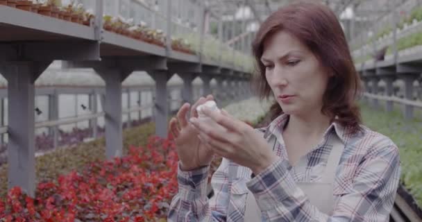 Retrato de mujer enfocada leyendo etiqueta en botella blanca con pesticidas. Bióloga agrónoma caucásica adulta que examina fertilizante en invernadero. Cultivo, jardinería. Sede del cine 4k ProRes — Vídeo de stock