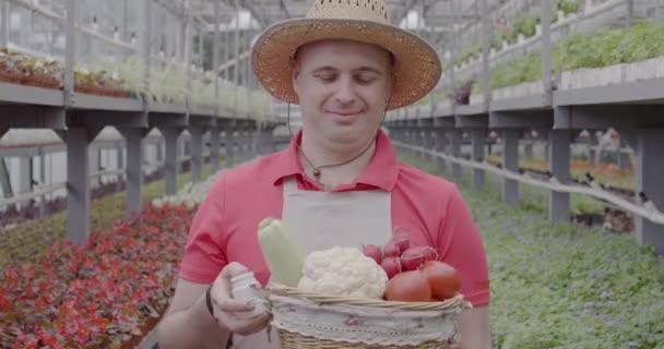 Lächelnder Mann mit Gemüsekorb, der eine Flasche mit Pestiziden in die Kamera reckt. Porträt des selbstbewussten positiven kaukasischen Biologen Agronom mit Strohhut, der im Gewächshaus posiert. Cinema 4k ProRes Hauptquartier. — Stockvideo