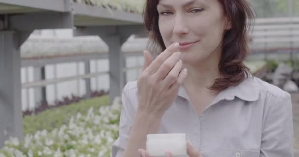 Cámara se mueve hacia arriba de la botella con crema hidratante orgánica a la cara de sonriente hermosa mujer caucásica oliendo y aplicando crema. Retrato de una encantadora dama en el invernadero. Sede del cine 4k ProRes . — Vídeo de stock