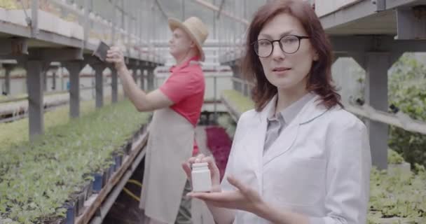 Профессиональная женщина-биолог в очках хвастается бутылкой удобрений перед камерой с довольным мужчиной-фермером, любующимся растениями на заднем плане. Садоводство, наука, теплица. Штаб-квартира кинотеатра 4k ProRes . — стоковое видео