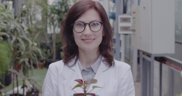 Primer plano de la mujer sonriente estirando pote a cámara. Retrato de mujer caucásica satisfecha con anteojos y bata médica mostrando planta en invernadero. Sede del cine 4k ProRes . — Vídeo de stock