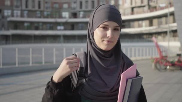 Vakker muslimsk kvinne i hijab som poserer med bøker utendørs på solskinnsdag. Portrett av sjarmerende student fra Midtøsten som smiler til kameraet. Utdanning, livsstil, innvandrere. – stockvideo