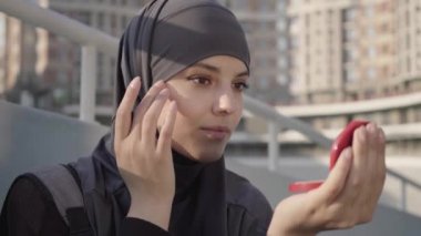 Açık havada yüz pudrası süren tesettürlü çekici Orta Doğulu genç bir kadın. Güzel müslüman bir kızın portresi. El aynasına bakıyor ve yüzüne dokunuyor. Makyaj, güzellik, yaşam tarzı.