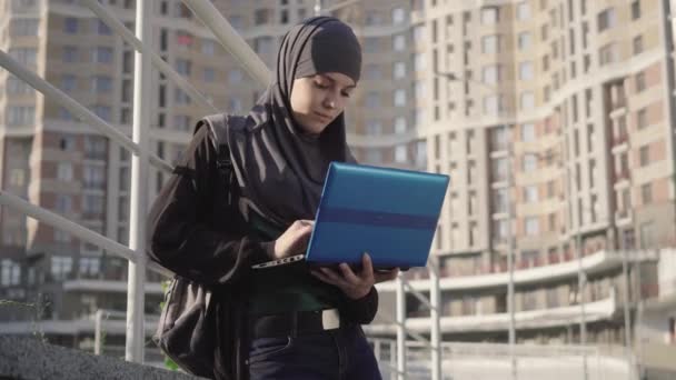 Geleneksel tesettürlü gülümseyen Müslüman kadın dışarıda dizüstü bilgisayarda yazı yazıyor. Şehir caddesinde sosyal medyayı kullanan güzel bir Orta Doğulu genç kız. Yaşam tarzı, kablosuz iletişim, çeşitlilik. — Stok video