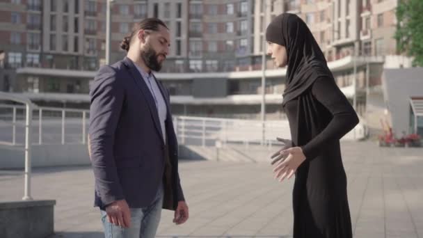 Boze jonge moslim vrouw schreeuwt naar een bebaarde, zelfverzekerde man uit het Midden-Oosten. Zijaanzicht van man en vrouw die buiten ruziën op zonnige dag. Jaloezie concept, relatieproblemen, conflict. — Stockvideo