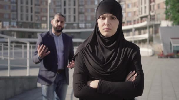 Retrato de una mujer triste en hiyab de pie con las manos cruzadas y mirando a la cámara como un hombre enojado gritando al fondo. Joven esposa de Oriente Medio con los ojos adoloridos y marido furioso peleando . — Vídeos de Stock