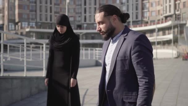 Öfkeli Orta Doğulu bir adamın arka plandaki tesettürlü Müslüman kadına bakıp kızgın bakışlarını kameraya çevirmesinin ortası. Zorba karı-koca portresi. Şiddet. — Stok video