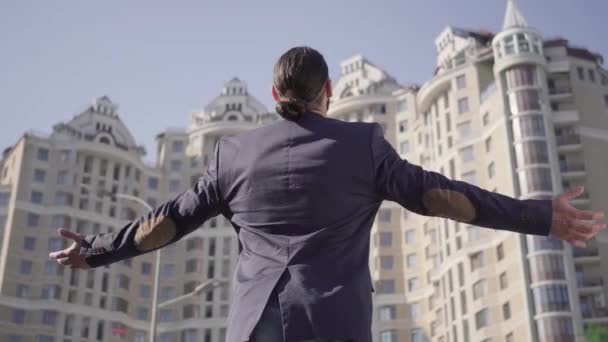 Вид на взрослого человека с Ближнего Востока с черным хвостиком, поднимающим руки и жестом. Успешный бизнесмен в костюме стоит перед городским небоскребом. Недвижимость, богатство, богатство . — стоковое видео