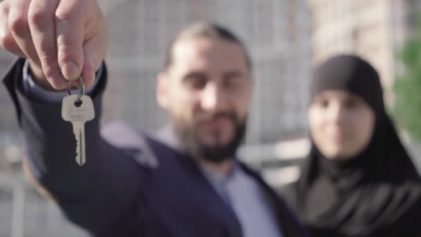 Detailní záběr klíče v mužské ruce s rozmazaným párem z Blízkého východu, usmívající se na pozadí. Šťastní majitelé nemovitostí chlubit vlastnictví nemovitostí venku za slunečného dne. Bohatství, úspěch, životní styl.. — Stock video