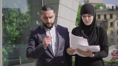 Başörtüsü içinde meşgul genç bir kadının portresi ve şehir caddesinde yürüyen ve belgeleri tartışan konsantre olmuş Orta Doğulu bir adam. Erkek ve kadın girişimciler güneşli bir günde dışarıda takım çalışmasını tartışıyor..