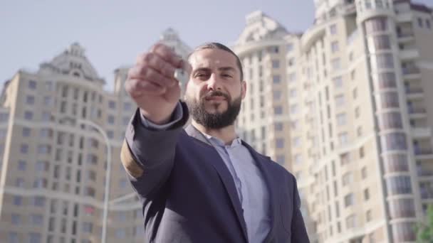 肯定的な成功した中東のビジネスマンはキーを投げ、キャッチし、勝利のジェスチャーを作る。都会の超高層ビルを背景にした幸せな豊かな不動産所有者の肖像画。ライフスタイル. — ストック動画