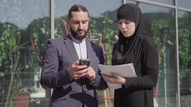 Mellanslag av självsäker muslimsk kvinna med dokument och man med smartphone diskuterar affärer utomhus. Porträtt av Mellanösterns folk som pratar på stadsgatan. Affärslivsstil. — Stockvideo