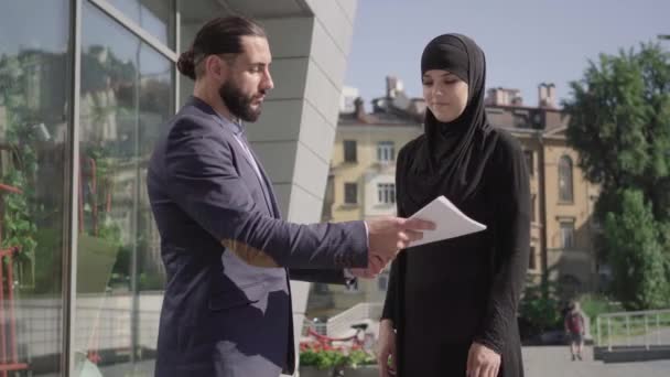 Позитивно улыбающиеся ближневосточные мужчина и женщина пожимают руки на улице после подписания деловой сделки. Рукопожатие уверенной мусульманки в хиджабе и бородатый мужчина в костюме. Командная работа, сотрудничество . — стоковое видео
