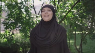 Neşeli genç Müslüman kadın alışveriş torbalarıyla el kaldırıyor ve kameraya gülümsüyor. Güneşli parkta açık havada poz veren tesettürlü mutlu Orta Doğulu kız portresi. Alışveriş çılgınlığı..