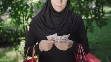 Banknotları sayıp gülümseyen tesettürlü tanınmamış bir kadın. Alışveriş torbalarıyla alışveriş yapan genç ve mutlu bir Orta Doğulu. Moda, alışveriş, yaşam tarzı, mutluluk, başarı..