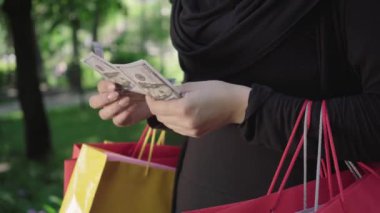 Güneşli bir yaz gününde, ellerinde alışveriş torbalarıyla parktaki dolar banknotlarını sayan kadınların yakın çekimleri. Geleneksel giysiler içinde tanınmayan Müslüman bir kadın satın aldıktan sonra elinde para tutuyor. Alışveriş çılgınlığı..