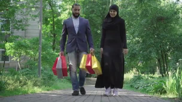 Heureuse femme musulmane en hijab et homme barbu confiant en costume approchant de la caméra et partant. Portrait large du riche couple moyen-oriental marchant dans un parc d'été ensoleillé avec des sacs à provisions. — Video