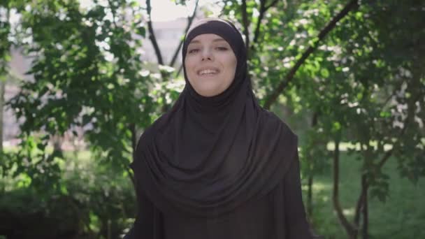 Joven mujer musulmana alegre levantando las manos con bolsas de compras y sonriendo a la cámara. Retrato de la feliz chica de Oriente Medio en hijab satisfecho con las compras posando al aire libre en el soleado parque. Shopaholism . — Vídeos de Stock