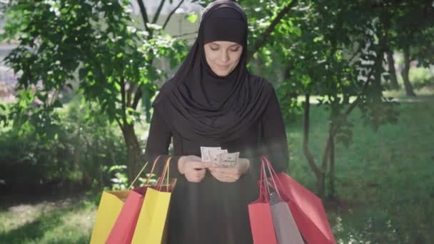 Sorrindo mulher feliz em hijab de pé em raios de sol com sacos de compras e pacote de dólares. Retrato de jovem alegre Oriente Médio menina satisfeita com compras na sexta-feira preta. Estilo de vida, moda . — Vídeo de Stock