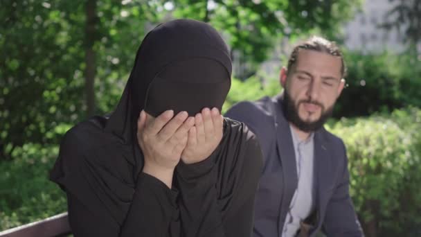 Gros plan d'une femme musulmane en hijab pleurant assise sur un banc alors qu'un homme du Moyen-Orient lui parlait de fond en comble. Mari calmant femme dans un parc d'été ensoleillé. Empathie, soutien, amour. — Video